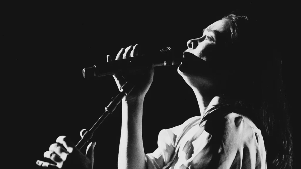Mitski singing into a handheld microphone, photographed in high-contrast black and white