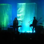 Beach House performing in silhouette against green and blue lit fabric columns