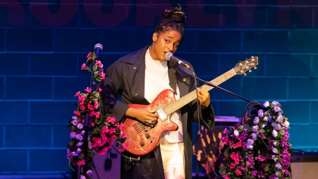 Taja Cheek of L'Rain singing on stage with a pink electric guitar and flower-wrapped microphone stands