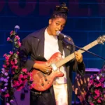 Taja Cheek of L'Rain singing on stage with a pink electric guitar and flower-wrapped microphone stands