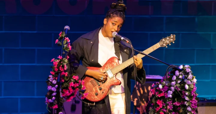 Taja Cheek of L'Rain singing on stage with a pink electric guitar and flower-wrapped microphone stands